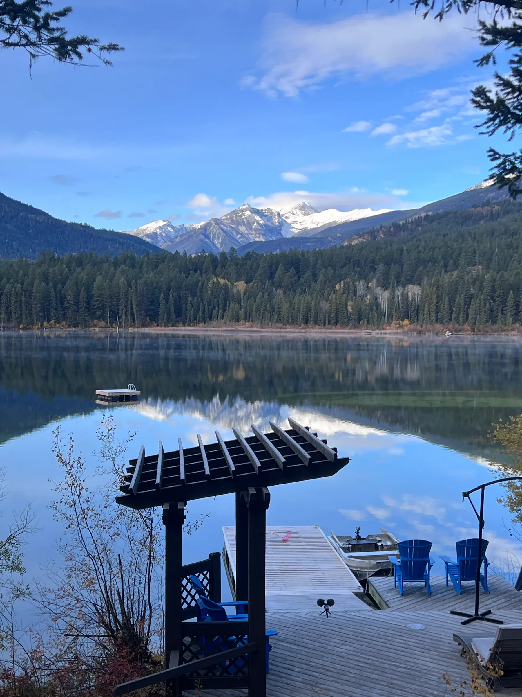A wooden dock with blue chairs and a pergola extends into calm water, reflecting snow-capped mountains and evergreen forests under a blue sky.